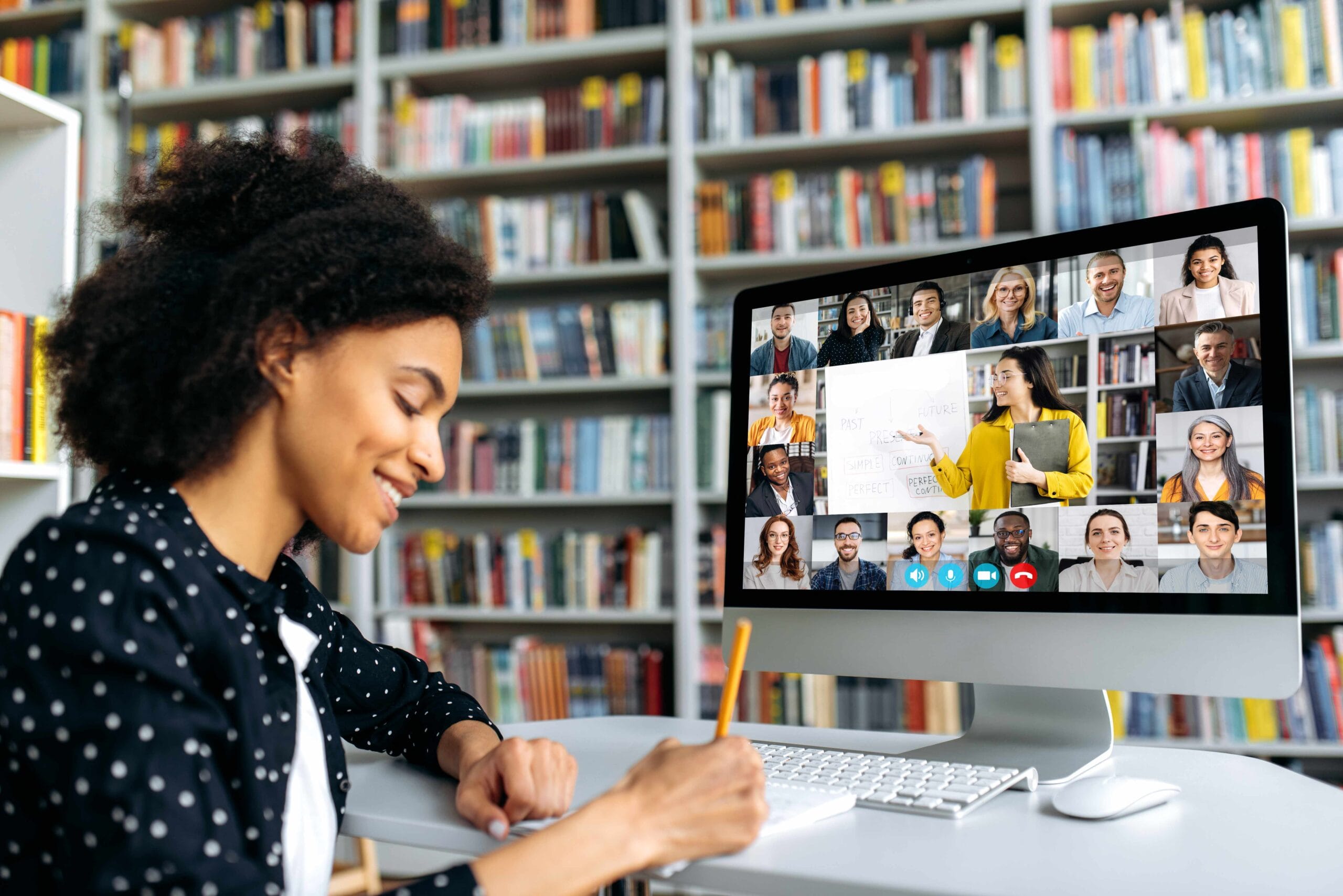 View over shoulder of a mixed race clever female student at a computer screen with a female teacher and students. Teacher conducts online lecture, female student takes notes. Online training,video call